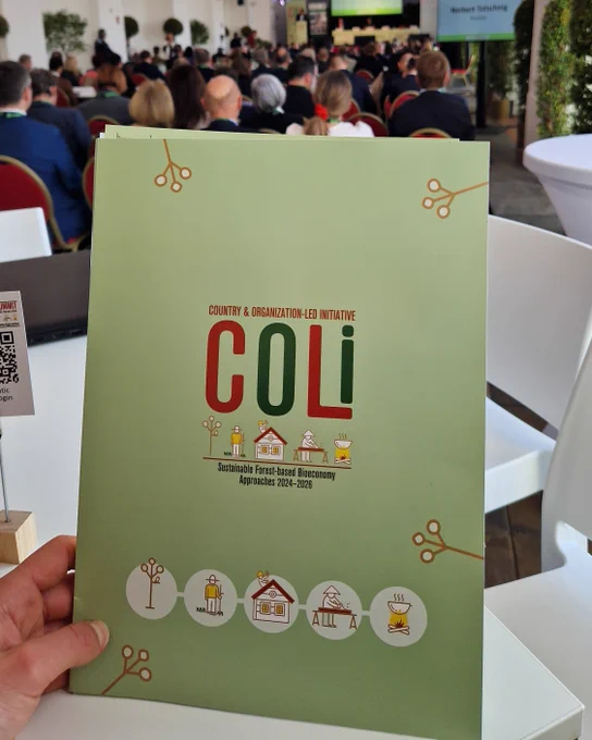 Hand holding a green booklet titled 'COLI' about sustainable forest-based bioeconomy approaches at the Vienna COLI summit with attendees in the background.