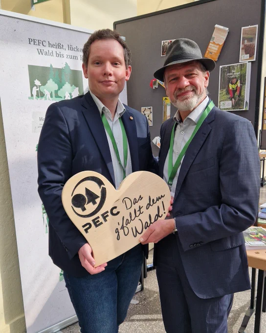 Two PEFC representatives holding a wooden heart-shaped sign with PEFC logo and German text promoting forest sustainability at the Vienna COLI summit.