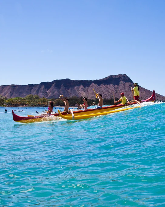 A group of people paddles a traditional outrigger canoe in bright blue waters, with Diamond Head crater visible in the background under a clear sky.