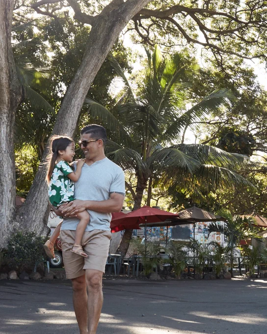 A person carrying a child is on a path surrounded by tall trees and greenery. The child is wearing a green and white outfit, and food stalls with red umbrellas are visible in the background. The atmosphere is sunny and warm.