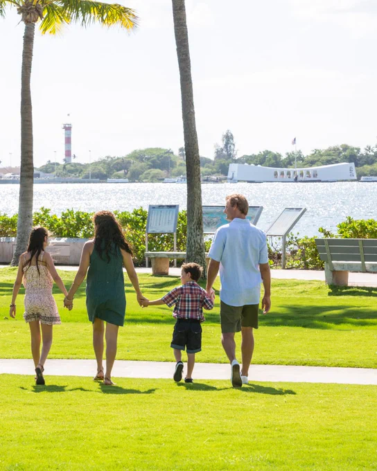 A group of four individuals walk hand in hand on a grassy path, with palm trees nearby and water in the background. The USS Arizona Memorial and a lighthouse are visible across the water.