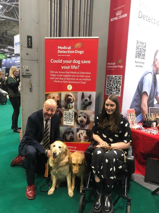 Steve Darling MP with Sophie and their assistance dogs in front of a sign which reads Medial detection dogs, could your dog save your life?
