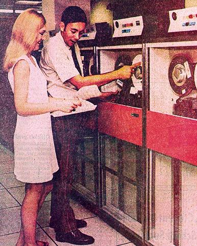 Two people operating vintage mainframe computers with large tape reels in a retro computer room setting.