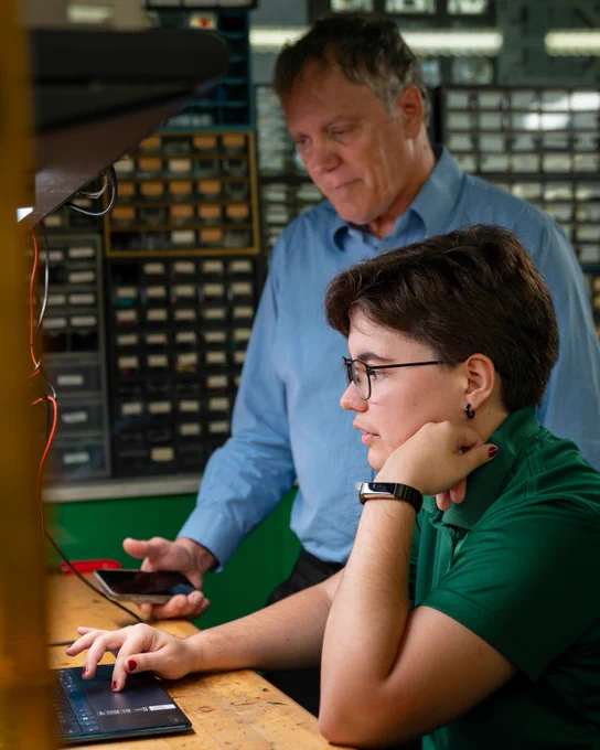 Two people engaged in a technical task at a workbench surrounded by electronic parts and equipment.