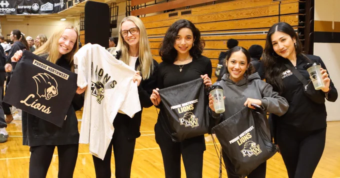 A group of five people stand indoors on a gymnasium floor, holding Purdue Northwest Lions merchandise, including shirts and bags. They are smiling and appear to be enjoying an event.