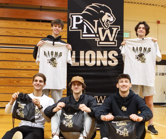 A group of five people is in a gymnasium, posing with t-shirts and drawstring bags featuring the Purdue Northwest (PNW) Lions logo. They are positioned around a PNW Lions banner, with three sitting on the floor and two standing behind. The setting is a basketball court with wooden bleachers in the background.