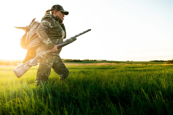 A person in camouflage clothing walks through a grassy field at sunrise or sunset, holding a shotgun and a turkey. The person wears a cap and is looking ahead. The landscape is expansive with a clear sky.
