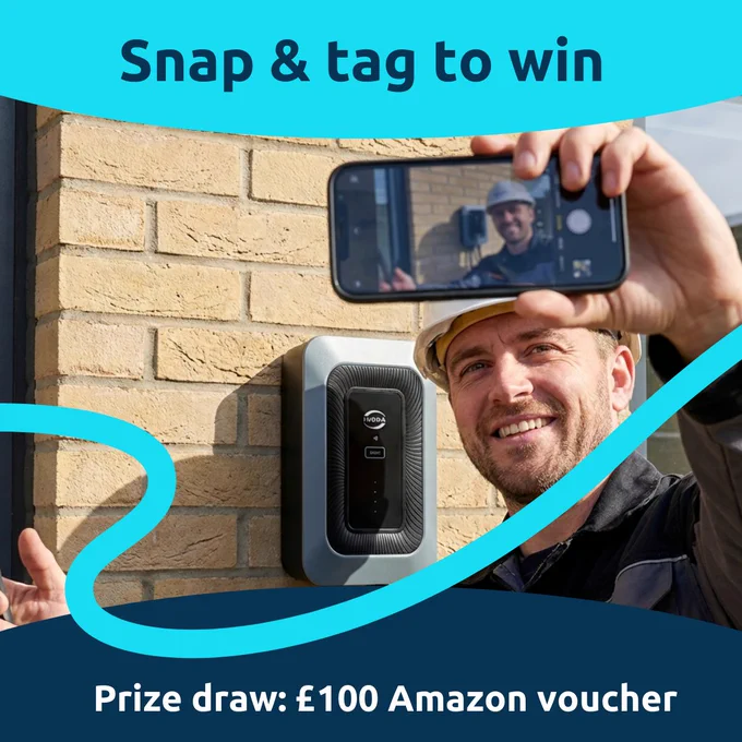A man stood in front of a home EV charger mounted on a brick wall holding up his phone to take a picture of himself and the charger.