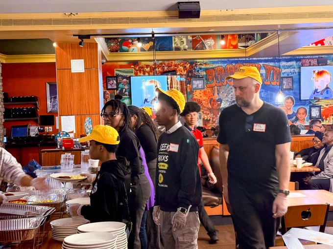A group of volunteers wearing yellow VISIONS caps stand in line to receive plates of breakfast. 