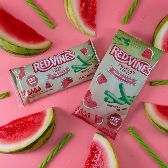 Packages of Red Vines Twisted Watermelon candy are placed on a pink surface, surrounded by slices of watermelon and green twisted candies.