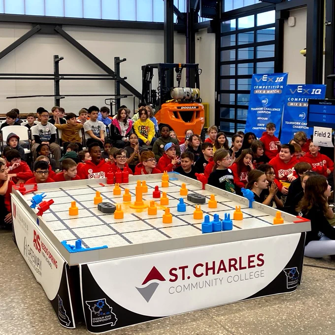Students gather around a robotic competition field featuring colorful game elements. The event is held at St. Charles Community College, with visible logos and a scoreboard in the background.