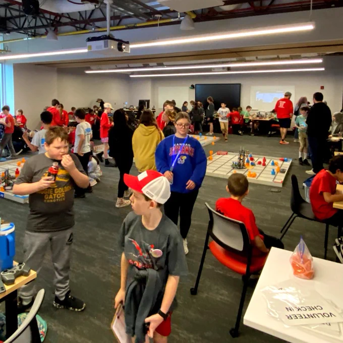 A room filled with people engaged in a robotics competition. Participants are gathered around tables with grids and various colored objects. Some individuals are observing, while others are actively participating. The atmosphere is lively and focused.