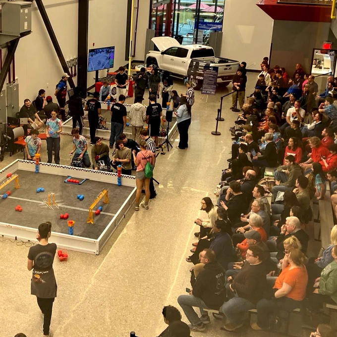 A robotics competition is taking place in a large indoor venue. Teams are gathered around a playing field with robots and various objects. A crowd of spectators is seated, watching the event attentively. A large display screen and a white vehicle are visible in the background.