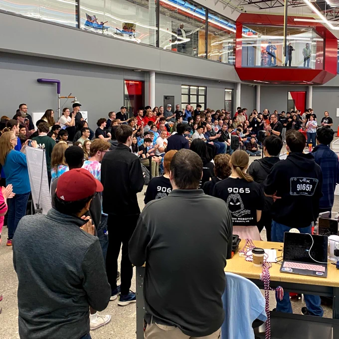 A large group of people are gathered at SCC's Regional Workforce Innovation Center in Wentzville for a robotics competition. Some individuals are wearing shirts with "9065" printed on the back. The setting is a modern facility with a balcony and large windows.