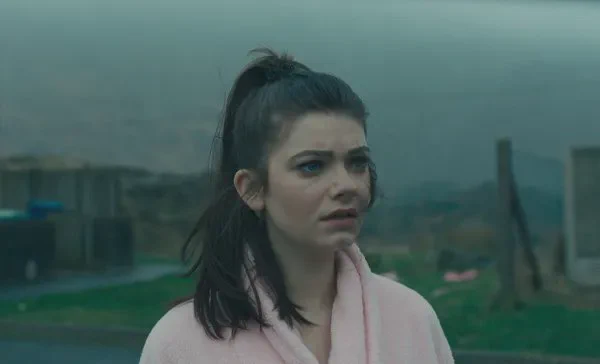 A still from the film Effi O Blaenau. A young white woman wearing a pale pink dressing gown stands in mist against a background of wasteland and dark mountain slopes.