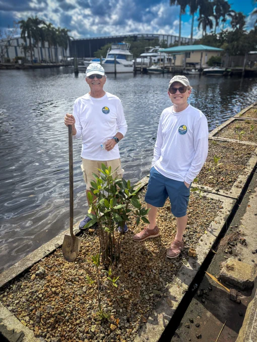 Two men in white long-sleeve shirts stand by a waterfront planting a small tree in rocky soil under a partly cloudy sky.