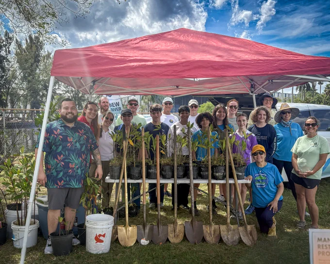 Group of volunteers standing under a red canopy with young plants and shovels ready for a tree planting event on a sunny day.