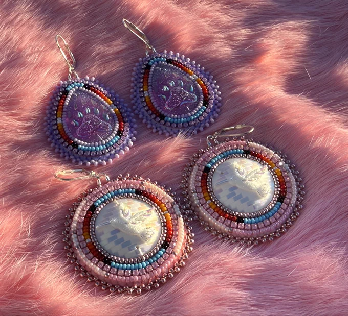 Native American Flat Stitched Beaded Earrings lying on pink rabbit fur. One pair is purple tear drop shaped with purple bear paw resin centers and the other is circular shaped with white resin hummingbird centers.