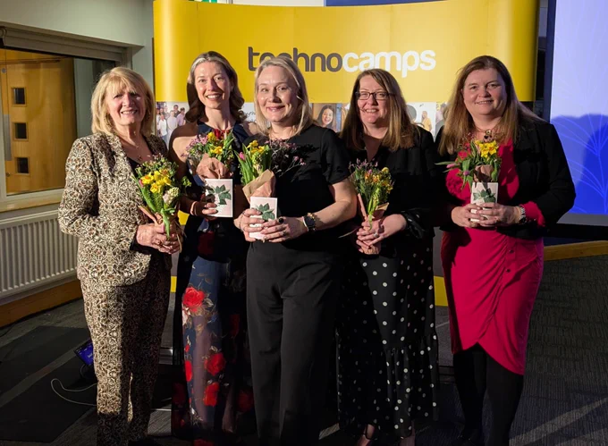 Sioned Williams MS standing with 4 women holding flowers in front of a Technocamps branded panel