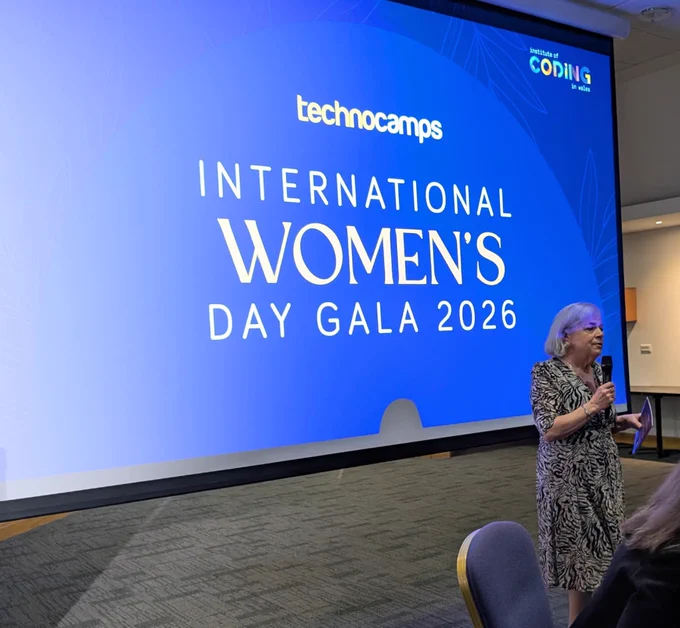 Beti Williams standing and speaking holding a microphone in front of a large floor to ceiling screen which has the text on it  “ Technocamps International Women’s Day Gala 2026”