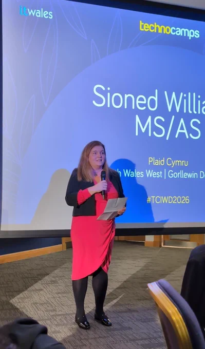 Sioned Williams MS standing and speaking holding a microphone in front of a screen which has her name on it and the text “ Plaid Cymru / South Wales West” and the hashtag #TCIWD2026