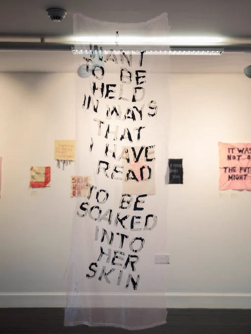 Translucent fabric hanging with black text reading 'I want to be held in ways that I have read to be soaked into her skin' in a gallery space.