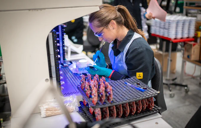 Technician wearing gloves inspects and works on electronic circuit boards under a UV light in a manufacturing facility.
