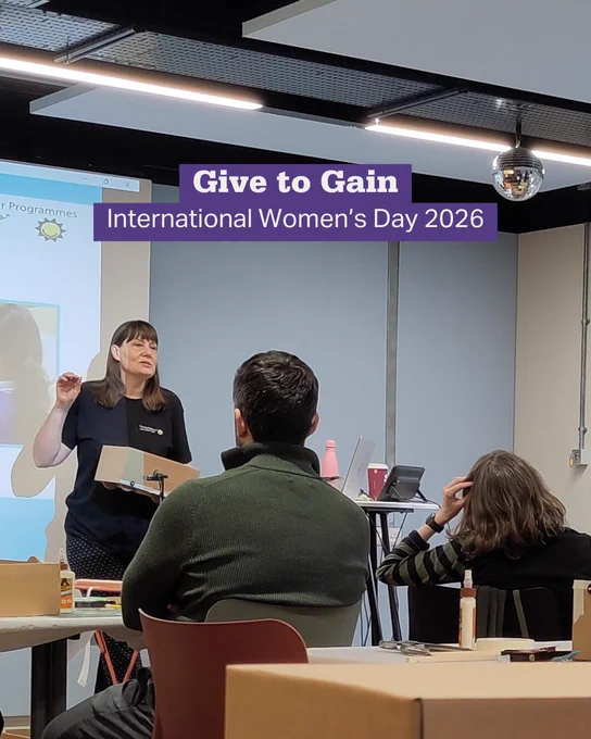 A woman presents at a seminar with a slide about International Women's Day 2026, titled 'Give to Gain'.