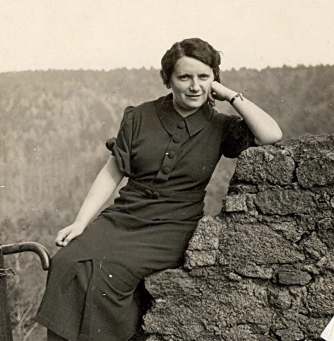 Vintage black and white photo of an woman sitting on a stone wall in a forested area, wearing a dark dress and resting their head on her hand.
