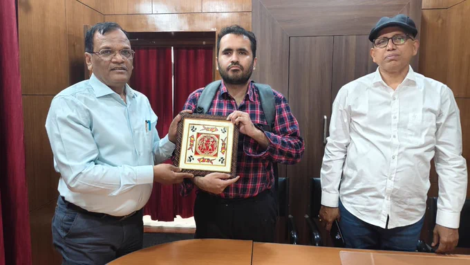 Three men standing in an office during a formal meeting, with one presenting a framed artwork to another, marking a discussion on accessibility and disability rights collaboration in Jharkhand.