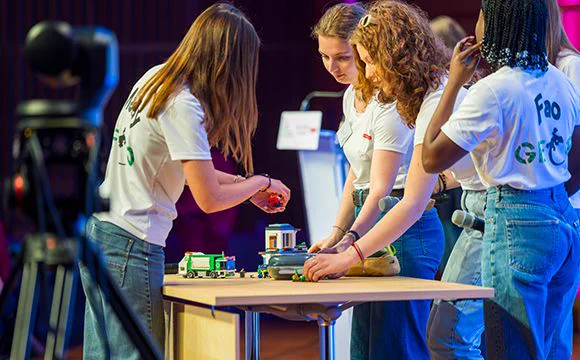 Quatre jeunes femmes, travaillent ensemble autour d'une table. Elles manipulent une petite construction type maquette ou robot (avec des briques et un petit véhicule vert). La scène semble se dérouler sur une scène ou lors d'un événement, avec un micro visible et du matériel en arrière plan. 