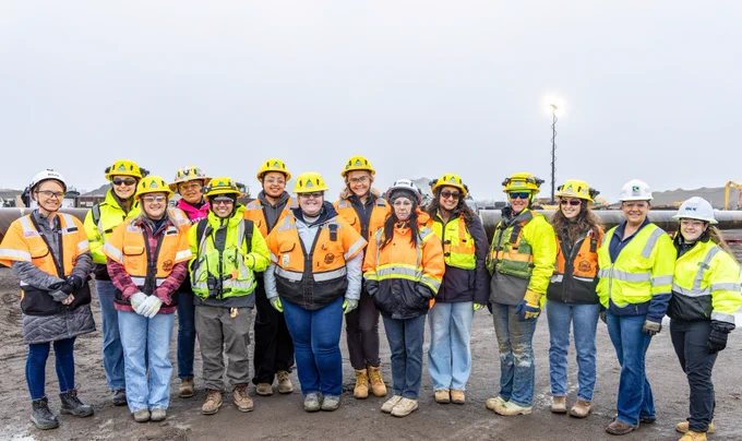 A group of construction workers wearing high-visibility jackets and hard hats stands together on a worksite. The ground is muddy, and the background features construction equipment and materials under an overcast sky.