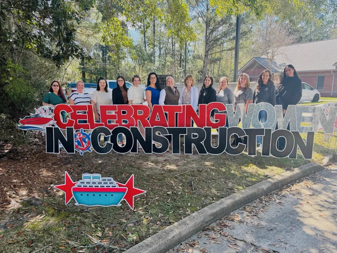 A group of individuals stands outdoors beside a large sign reading "Celebrating Women in Construction." Trees and a building are visible in the background.