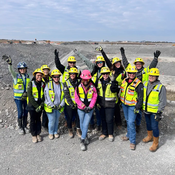 A group of construction workers wearing safety gear, including helmets and high-visibility vests, standing on a gravel area at a construction site. Some are raising their arms and smiling. The background shows piles of dirt and machinery.