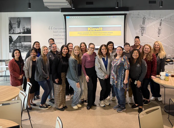 A group of people posing together in a room, in front of a large screen displaying "Kiewit Women in Construction Week." The background features architectural drawings and quotes on the wall. Tables and chairs are visible in the foreground.
