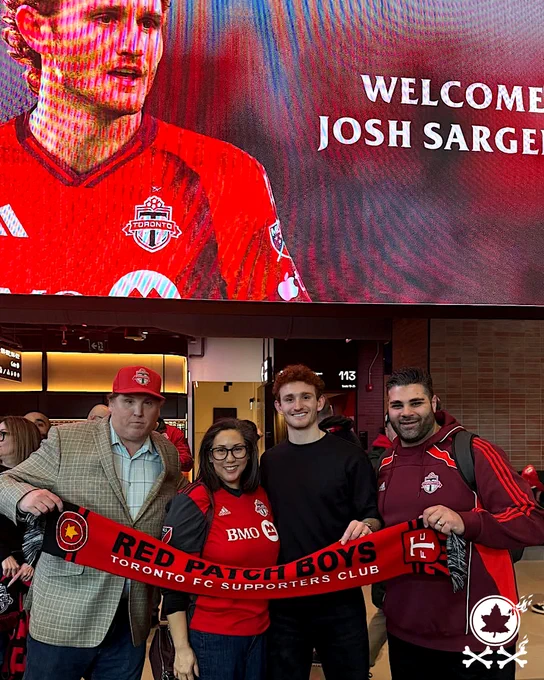 Red Patch Boys supporters group members standing with Josh Sargent holding a red scarf between them under a large illuminated TFC welcome screen with a picture of him in Toronto FC kit and the words WELCOME JOSH SARGENT. (bottom right corner white Red Patch 20-year logo with double-crossbones XX) March 2026.