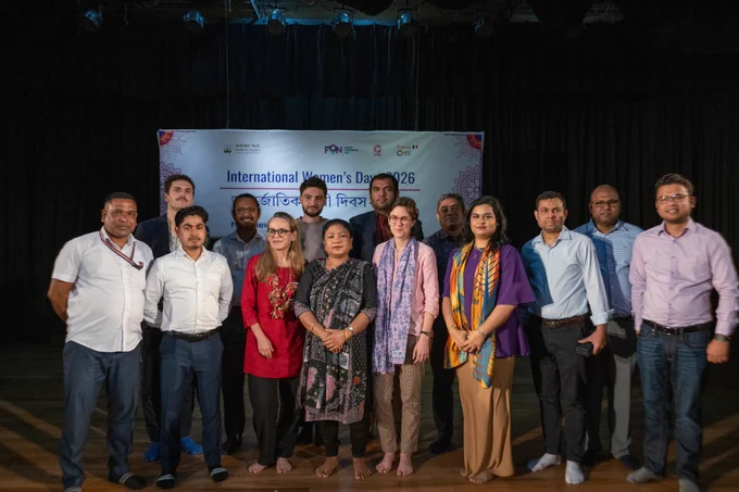 A mixed group of women and men stand together on stage for a group photo in front of an “International Women’s Day – 2026” banner. The group faces the camera under stage lighting at the event venue.
