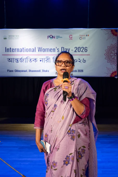 A woman in a light purple floral sari speaks into a handheld microphone on stage. She stands in front of a banner reading “International Women’s Day – 2026,” at the event held in Dhaka.
