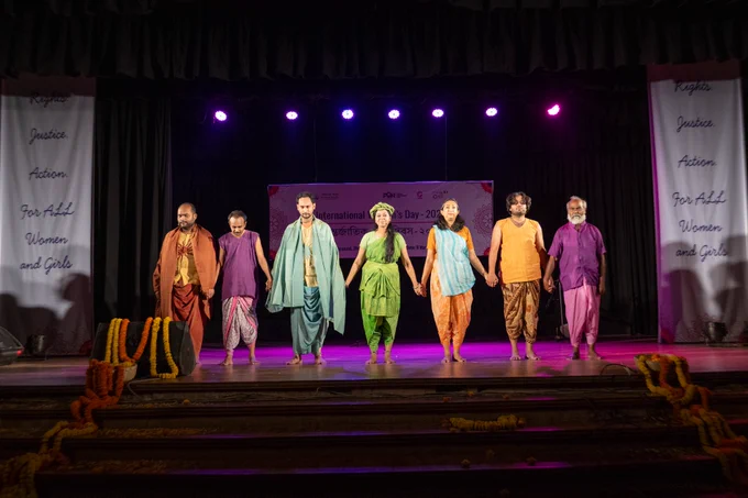 Seven performers stand barefoot in a line across a stage, holding hands during a performance under purple stage lights. Behind them is a banner for “International Women’s Day – 2026,” with side banners reading “Rights. Justice. Action. For All Women and Girls.”
