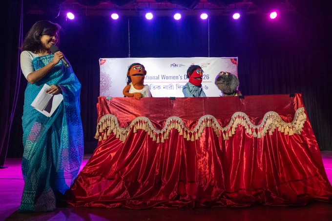 A host wearing a blue sari stands on stage holding a microphone and speaking while a puppet performance takes place beside her. Three puppets appear above a red curtain-covered puppet stage with gold fringe, set against the “International Women’s Day – 2026” banner.
