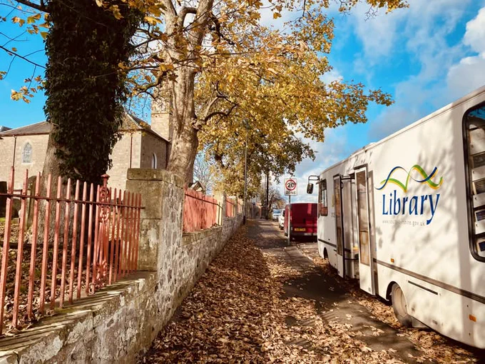 A mobile library vehicle is parked on a leaf‑covered pavement beside a stone wall and red metal fence. Large trees with golden autumn leaves line the street. A church-like stone building stands on the left, and another vehicle is parked ahead under a blue sky with scattered clouds.
