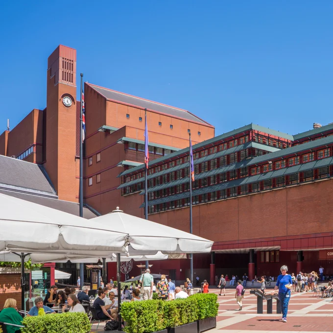 A view of the exterior of the British Library St Pancras building from the piazza, with people sitting in an outdoor cafe.