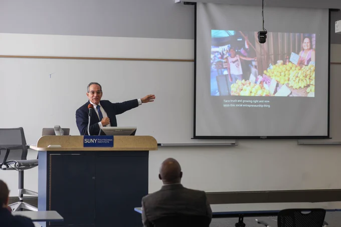 Wide view of a classroom at SUNY Polytechnic Institute as Dr. Martin Burt presents at the front, gesturing toward slides projected on two screens while audience members watch from seated tables.