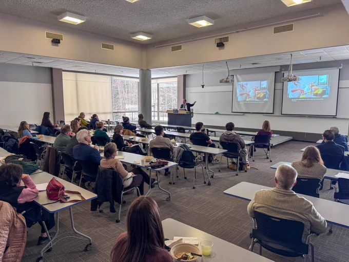 Dr. Martin Burt gestures toward a projected slide showing a market scene with fruit vendors while speaking at a SUNY Polytechnic Institute classroom podium.