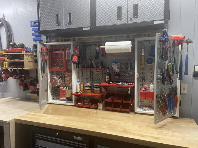 Wall-mounted cabinet workspace featuring Wall Control metal pegboard panels installed inside open cabinet doors and along the back wall, organizing hand tools, pliers, screwdrivers, hooks, shelves, red storage bins, and small hardware above a light wood workbench in a clean garage setting