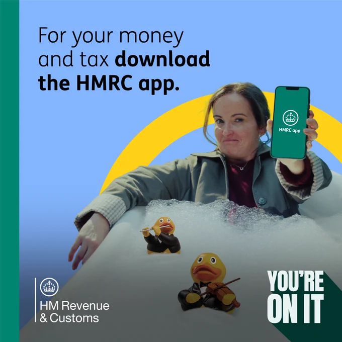 A fully dressed lady is sat in a bath of bubbles and two rubber ducks showing her phone with the HMRC app.