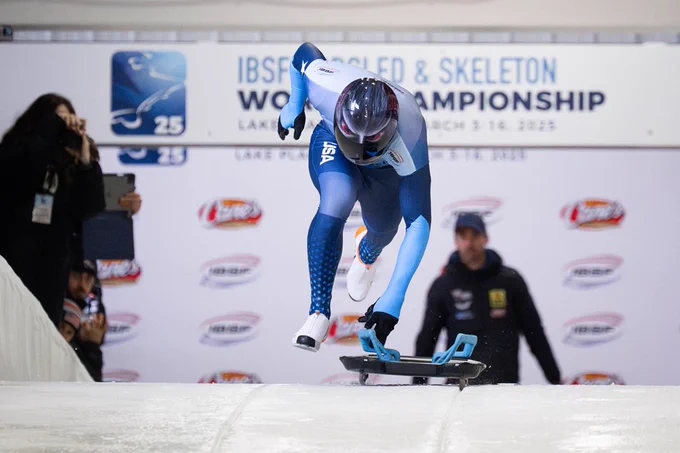 An athlete is starting a skeleton race at the IBSF Bobsled & Skeleton World Championship in Lake Placid. The athlete is wearing a USA uniform and helmet, and is pushing off on a sled. A banner and several logos are visible in the background.
Photo by James Reed Photography