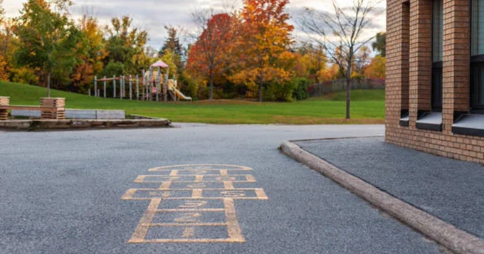 A sidewalk hopscotch grid is painted near a brick building, with a playground and autumn trees in the background.