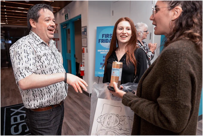 Three individuals engaging in a lively conversation at one of Barrie's Culture Night Out events, with one person holding a beverage and another holding a printed illustration. There are signs and banners in the background.