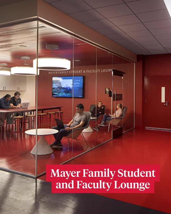 A modern lounge with large glass walls, labeled "Mayer Family Student and Faculty Lounge." Inside, several people are seated on stylish chairs, with some using laptops at tables. A large screen is visible on one wall. The lounge has bright red flooring and a sleek ceiling with circular light fixtures.